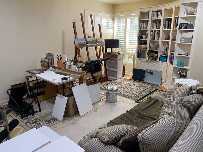 A photograph of the interior of the studio at our house. Bookshelves o the right, easel in the center and taboret /palette on the left side. There is a sofa in the foreground and everything looks slightly cluttered.