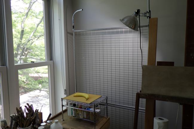 A photograph of part of an artist's studio with a still life set up at bottom center. There is a window on the left and an easel on the right.